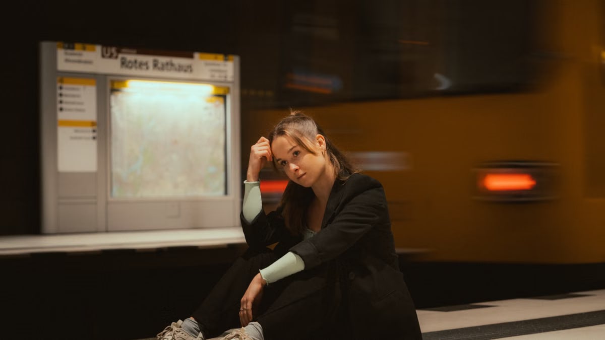 A young woman in casual attire seated at a Berlin subway station with trains in motion.